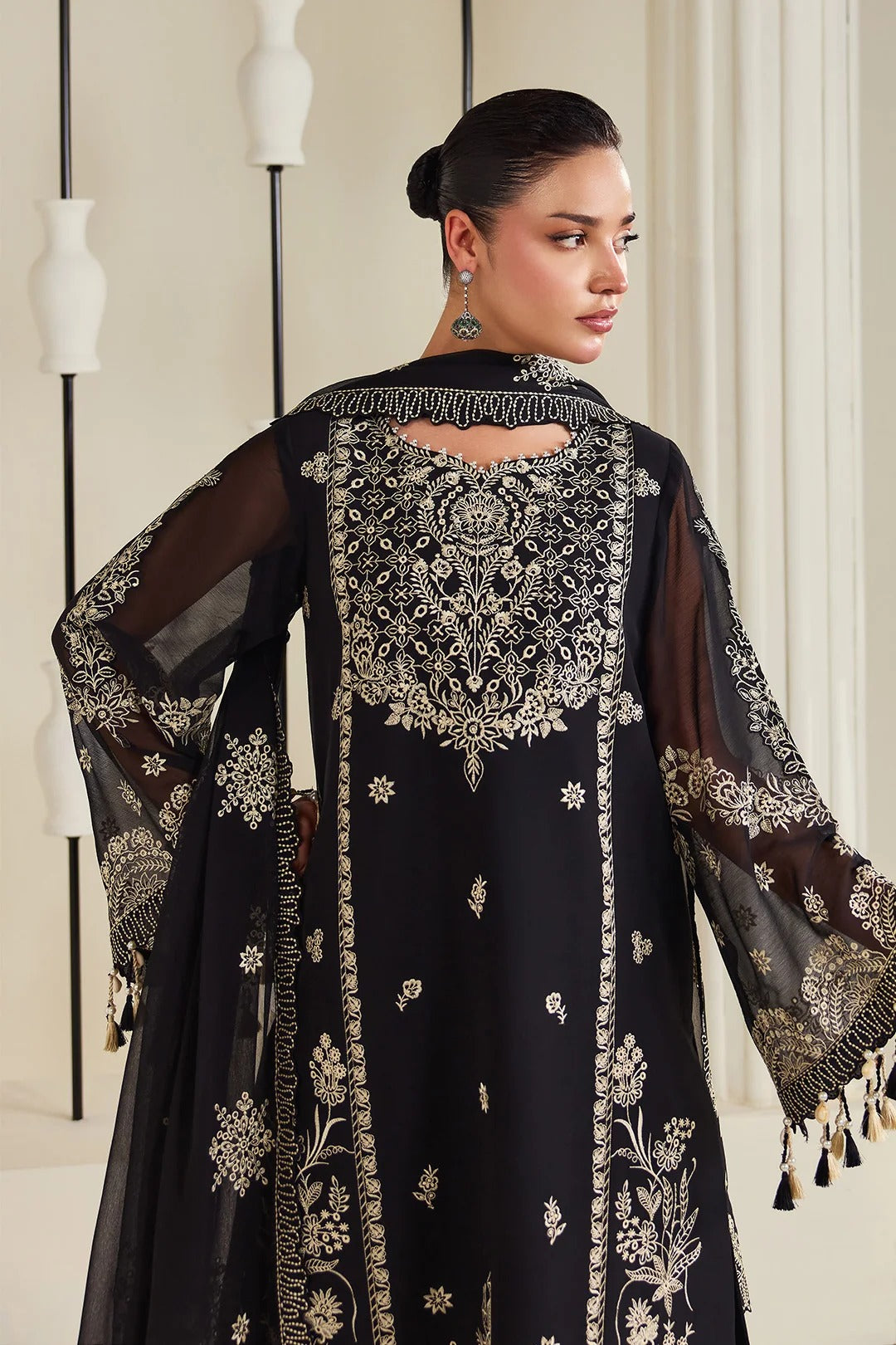 A woman modeling a black embroidered traditional outfit with sheer sleeves and ornate gold detailing, shown from the back in an indoor setting with hanging pendant lights.