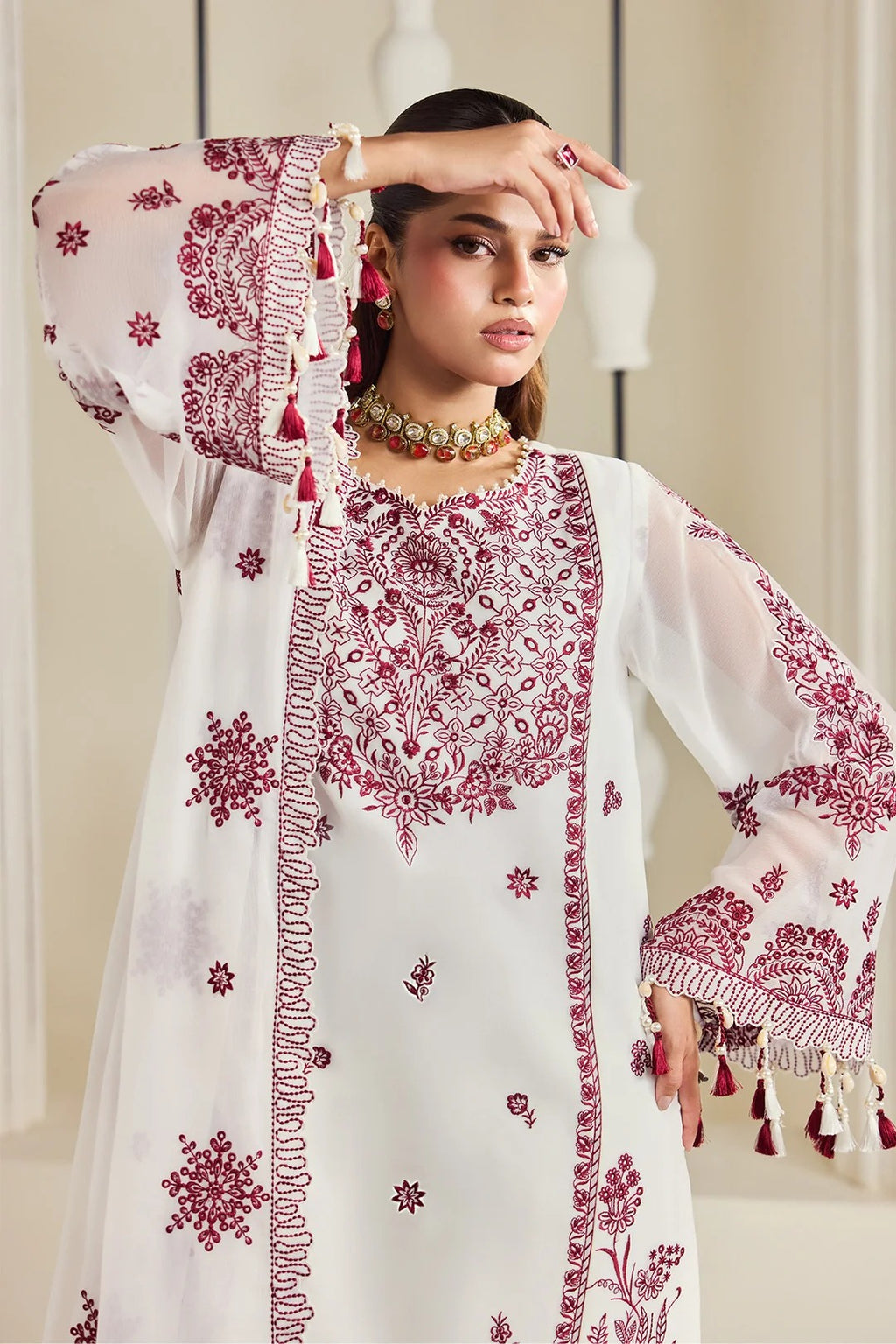 A woman models a white traditional embroidered kurta with deep maroon floral embroidery, tassel trim, and wide bell sleeves in an indoor setting.