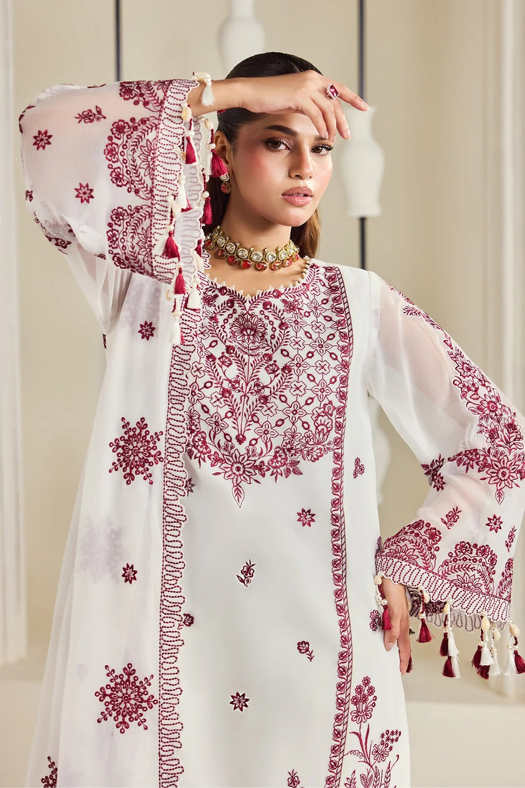A woman models a white traditional embroidered kurta with deep maroon floral embroidery, tassel trim, and wide bell sleeves in an indoor setting.