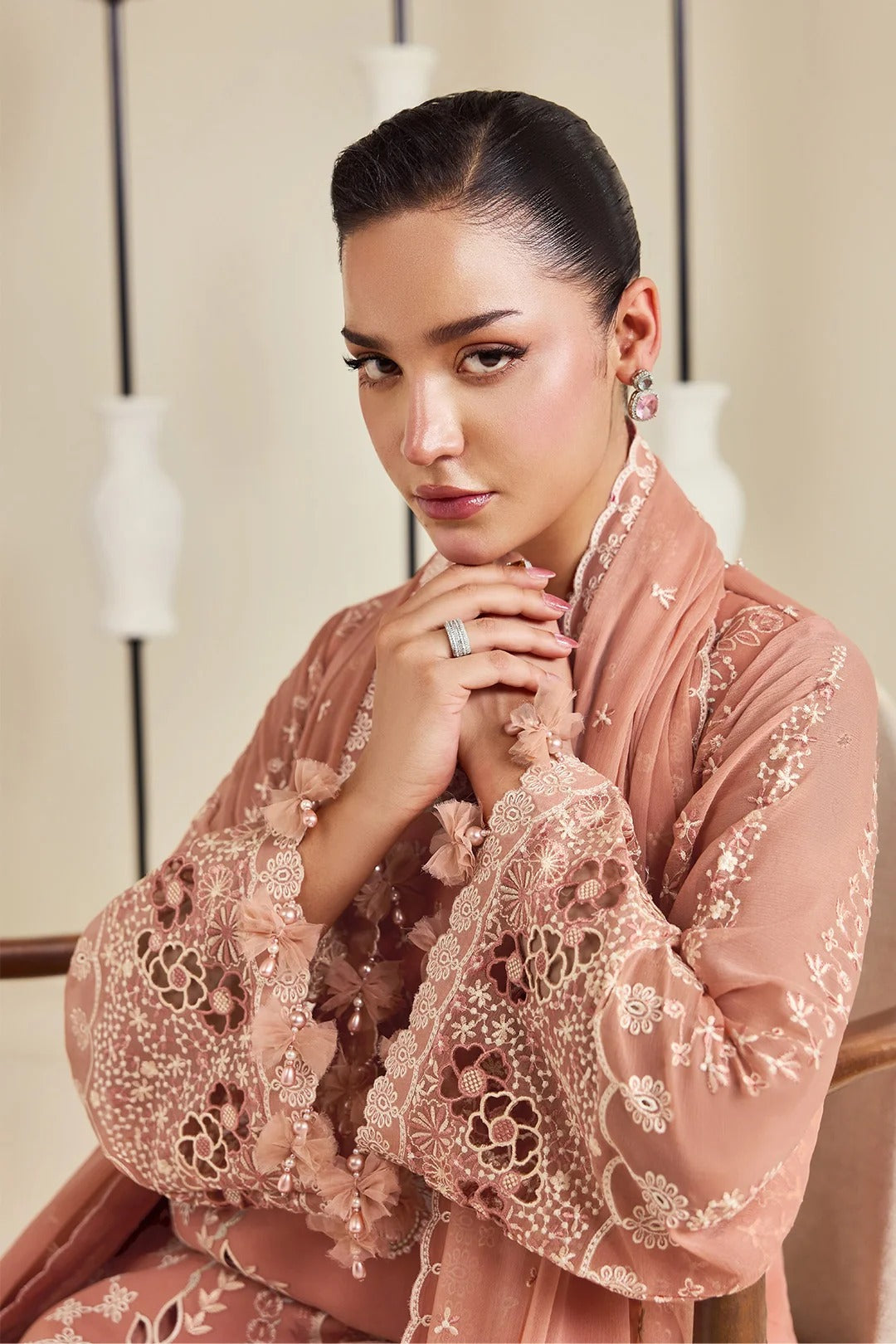 A woman wearing a dusty rose pink embroidered traditional outfit with intricate lace and floral detailing, posed in an elegant indoor setting.