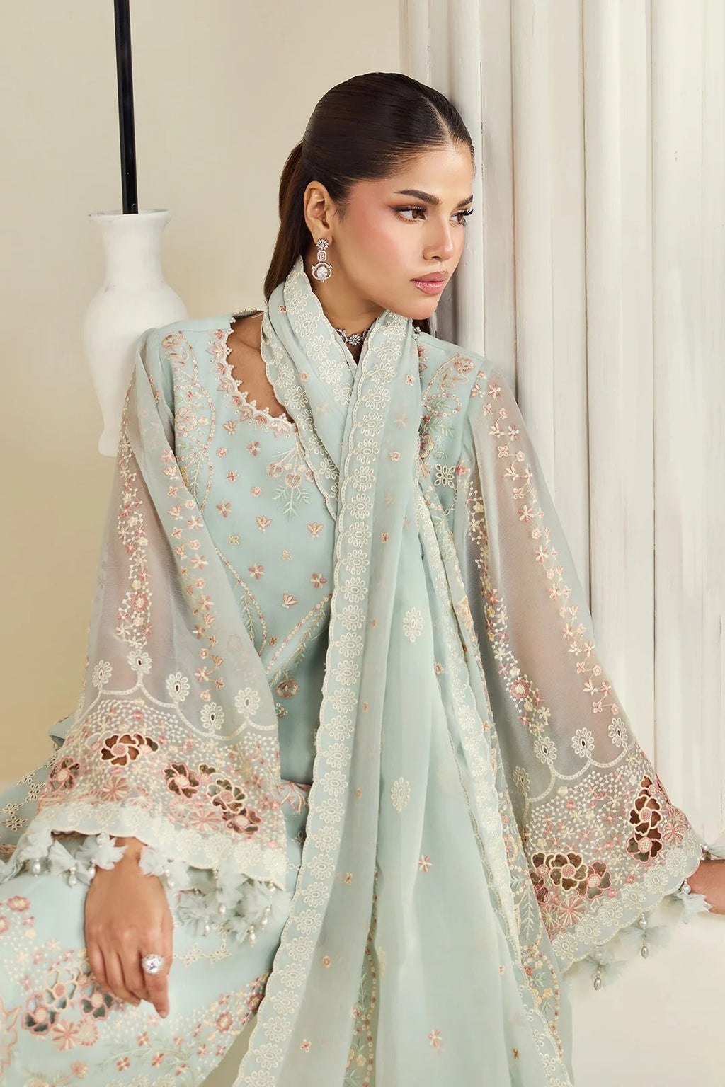 A woman models a traditional light blue-green ethnic outfit with matching sheer dupatta, featuring intricate floral embroidery and lace detailing in pink and beige, set against a neutral indoor background.