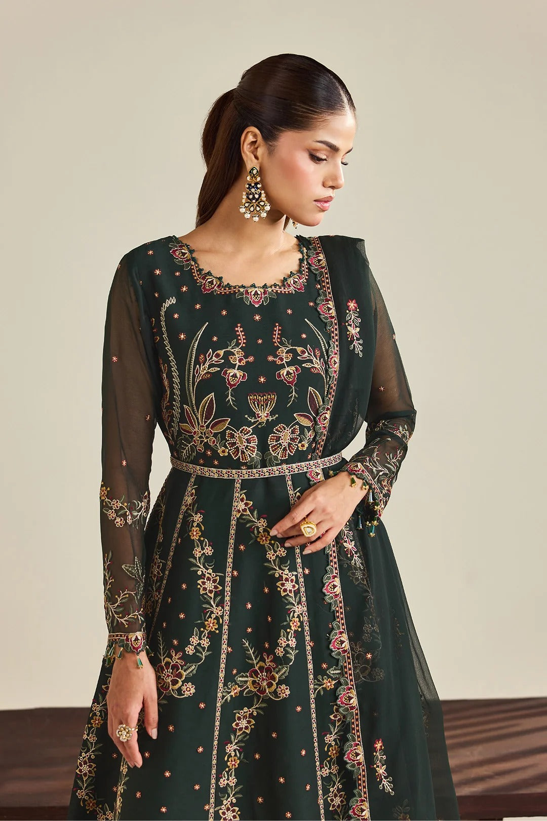 A woman models a dark green embroidered Anarkali gown with sheer long sleeves and intricate floral embroidery in gold, pink, and red, paired with a matching dupatta.