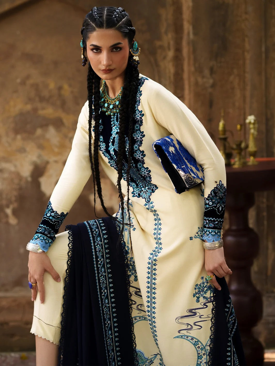 A woman models a cream-colored traditional South Asian outfit with intricate blue embroidery, paired with a dark navy dupatta and a blue clutch, set against a rustic stone wall background.