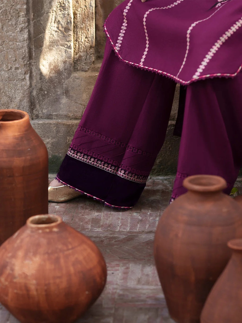 A close-up of a person wearing a deep purple traditional outfit with white embroidered details, standing among rustic terracotta clay pots against a stone wall.