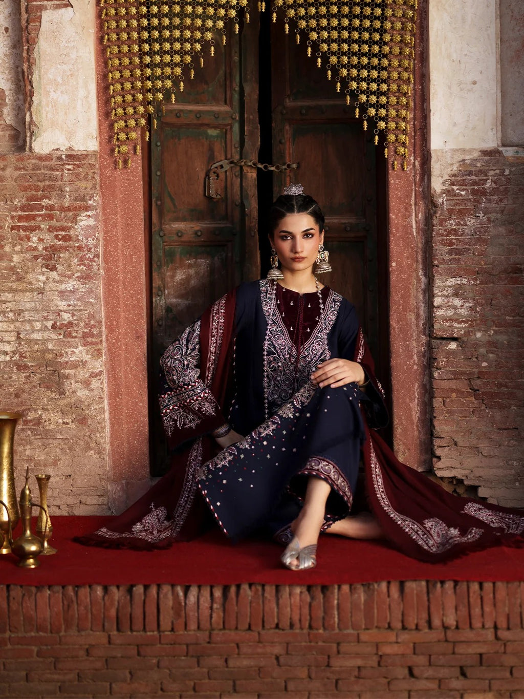 A woman sits on a red carpet in front of an ornate wooden doorway, wearing a dark navy and deep red embroidered traditional outfit with metallic embellishments and a matching draped fabric.