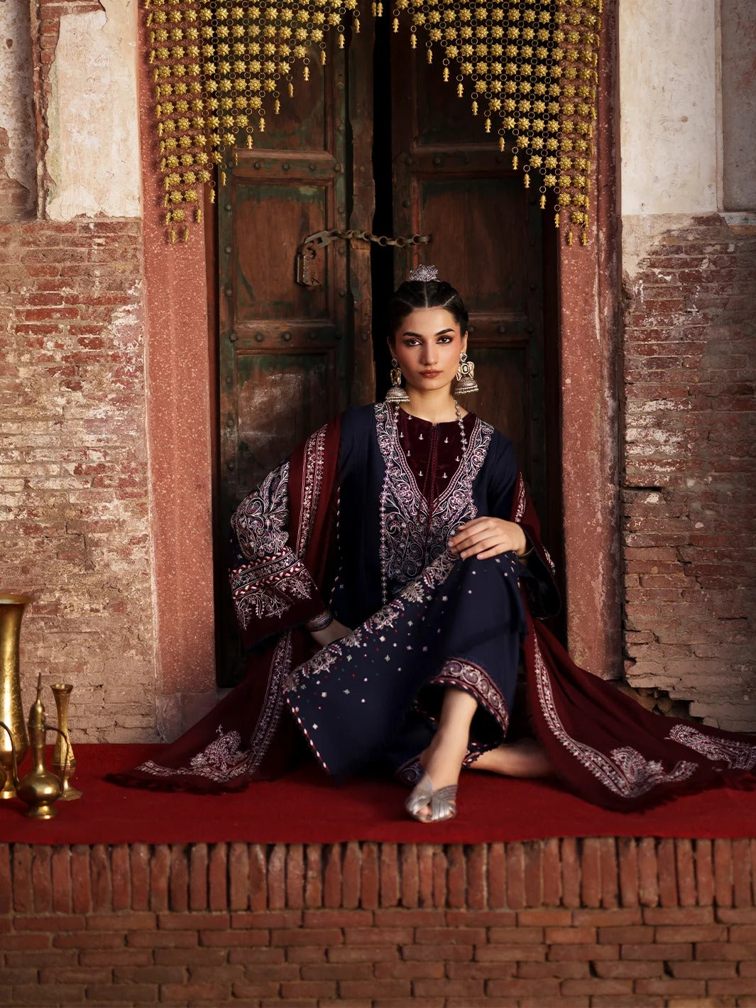 A woman sits on a red carpet in front of an ornate wooden doorway, wearing a dark navy and deep red embroidered traditional outfit with metallic embellishments and a matching draped fabric.