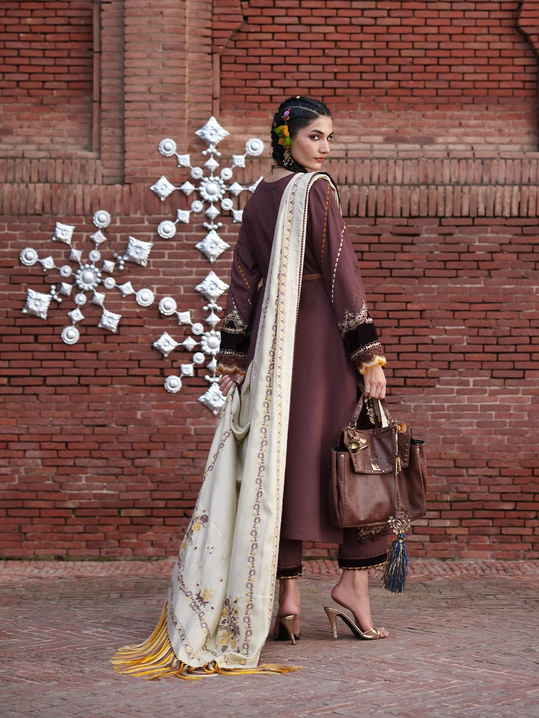 A woman models a traditional South Asian outfit consisting of a deep maroon kurta, matching trousers, and a cream-colored dupatta with gold embroidery and yellow fringe, set against a red brick wall background.