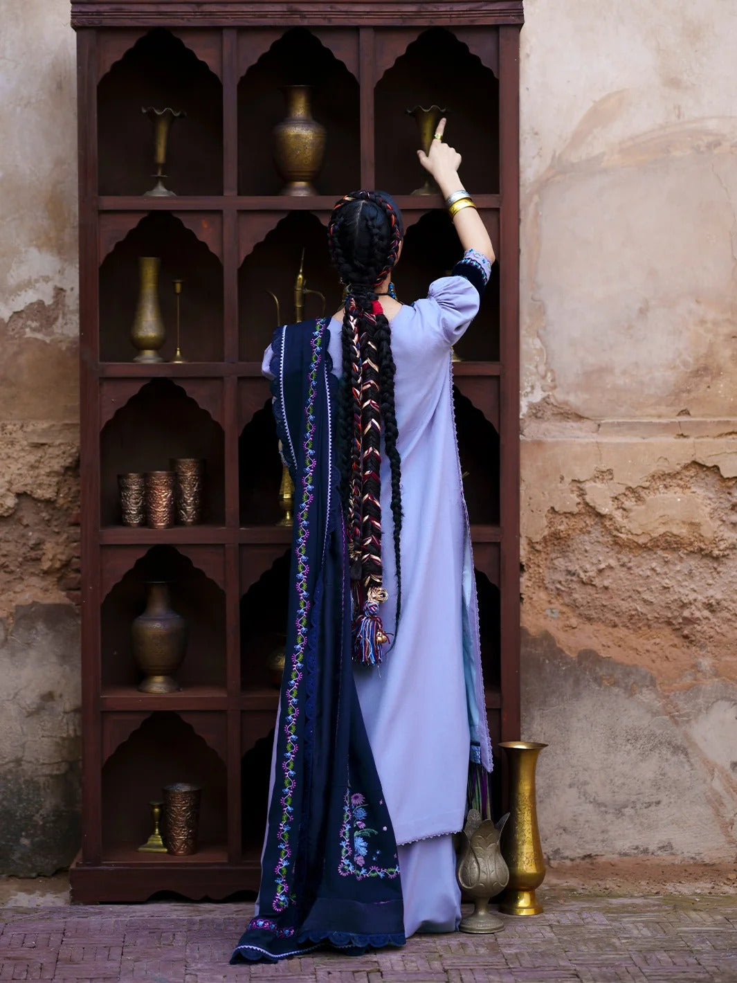 A woman viewed from behind wearing a traditional light blue and navy embroidered outfit, standing in front of a dark wooden display cabinet with brass vases against a textured beige wall.