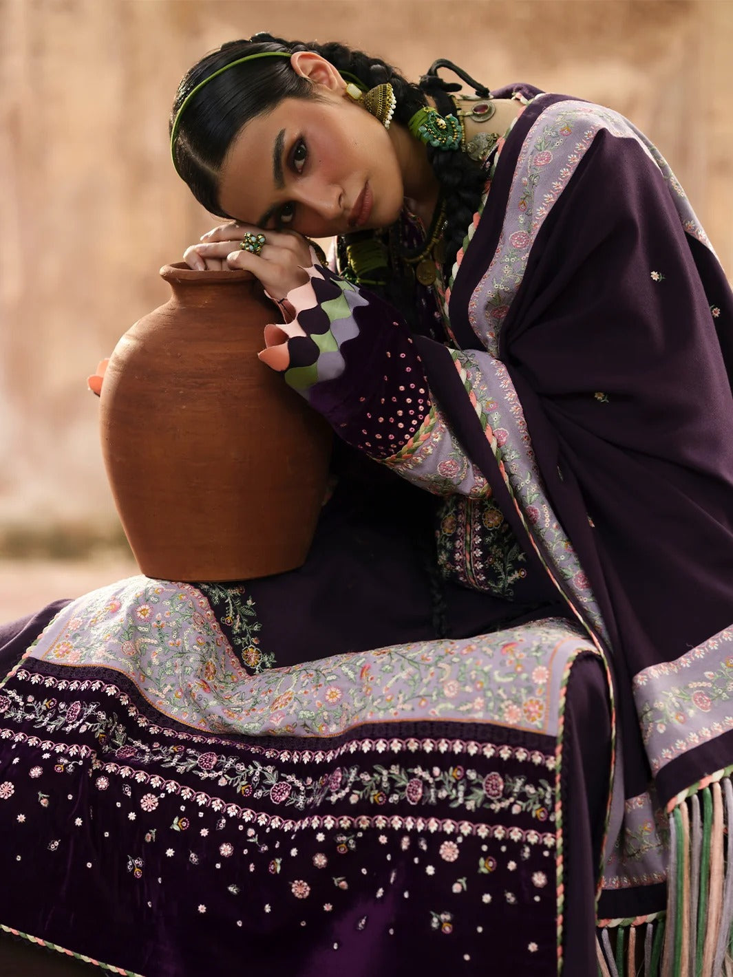 A woman in traditional South Asian attire wearing a deep purple embroidered outfit and matching dupatta, leaning against a large terracotta pot in a rustic setting.