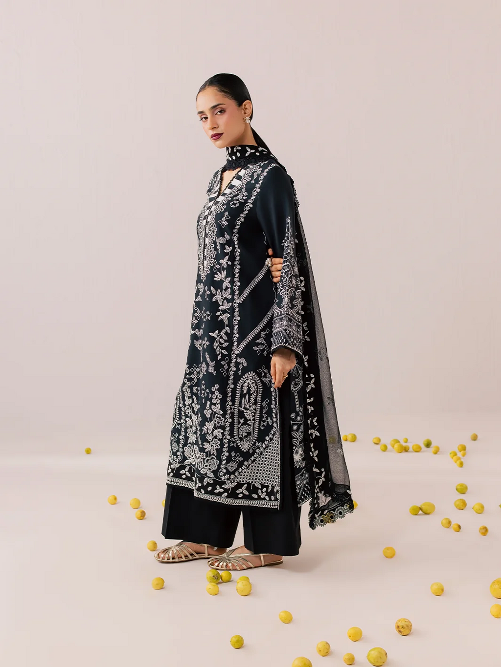 A woman models a black three-piece traditional outfit with intricate silver embroidery, featuring a long kurta, wide-leg pants, and a matching dupatta, set against a light beige background with scattered yellow pom-pom props.