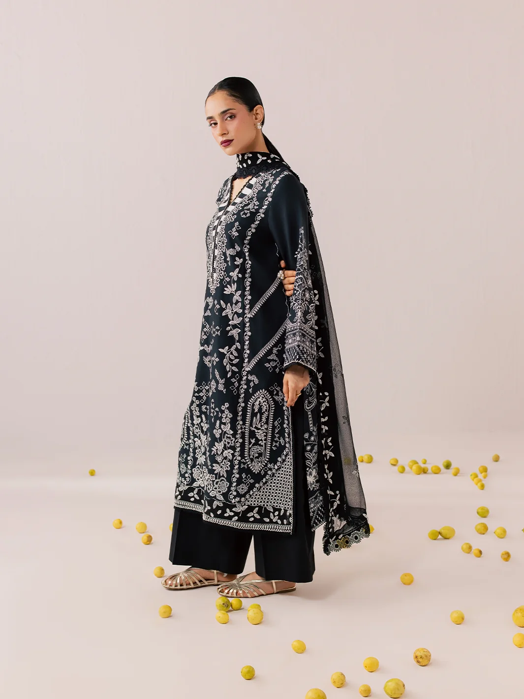 A woman models a black three-piece traditional outfit with intricate silver embroidery, featuring a long kurta, wide-leg pants, and a matching dupatta, set against a light beige background with scattered yellow pom-pom props.