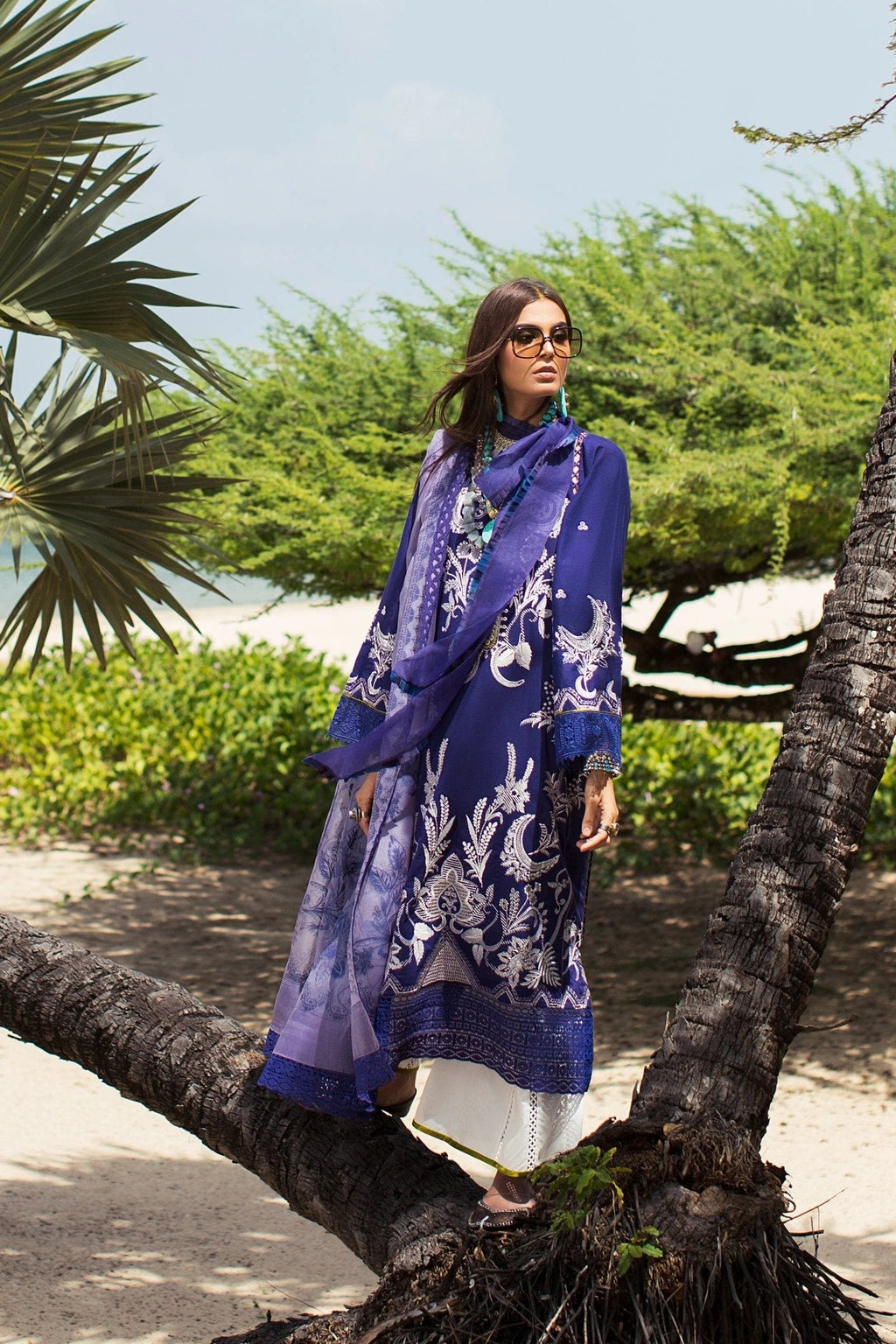 A woman wearing a royal blue printed ethnic kurta with a matching dupatta and white trousers, standing outdoors on a tree branch with palm trees and greenery in the background.