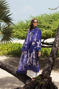 A woman wearing a royal blue printed ethnic kurta with a matching dupatta and white trousers, standing outdoors on a tree branch with palm trees and greenery in the background.