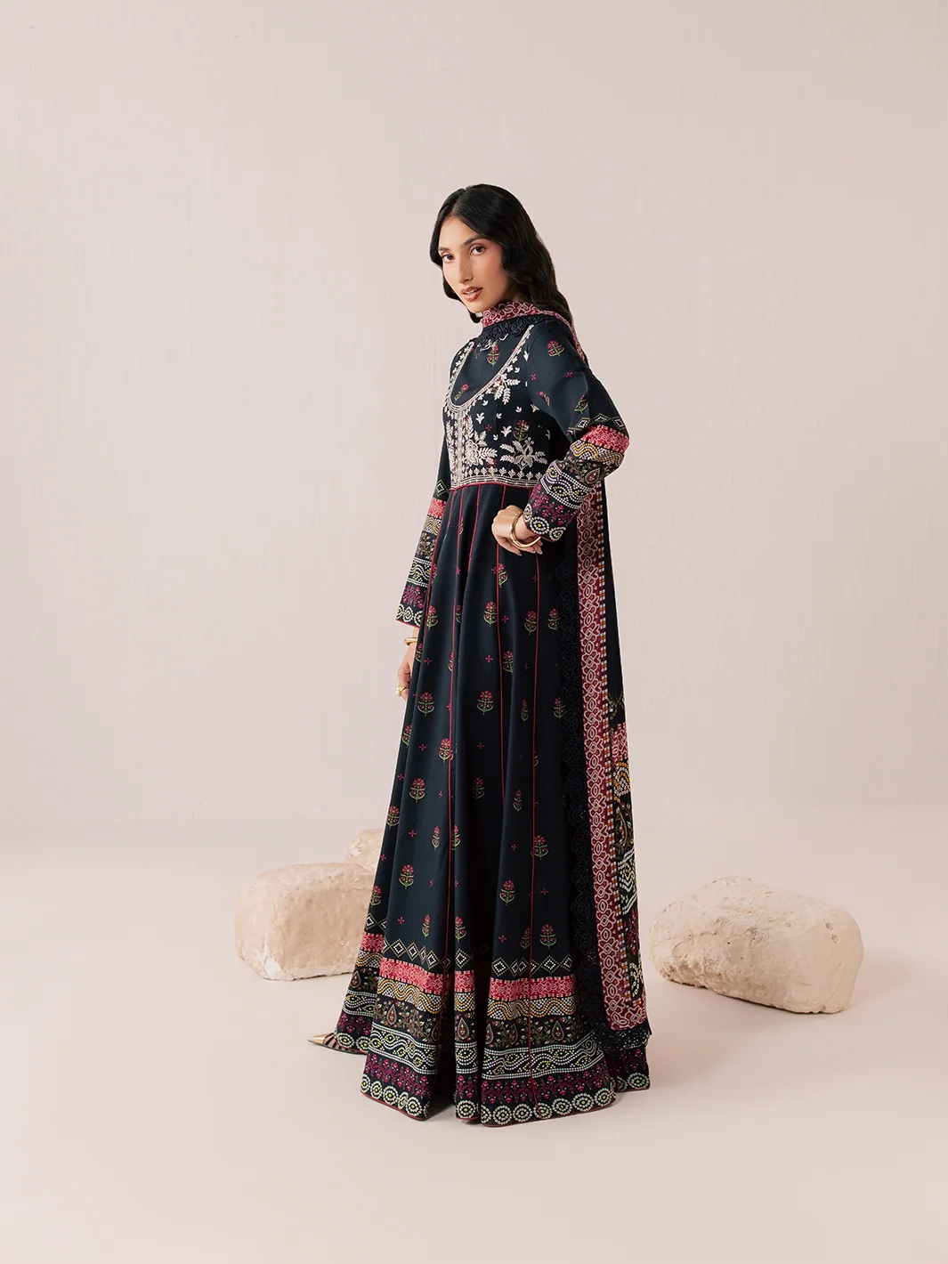 A woman models a dark navy blue traditional Anarkali gown with intricate silver embroidery on the bodice, pink and gold floral patterns, and a matching embroidered dupatta, set against a warm beige studio background with decorative stone props.