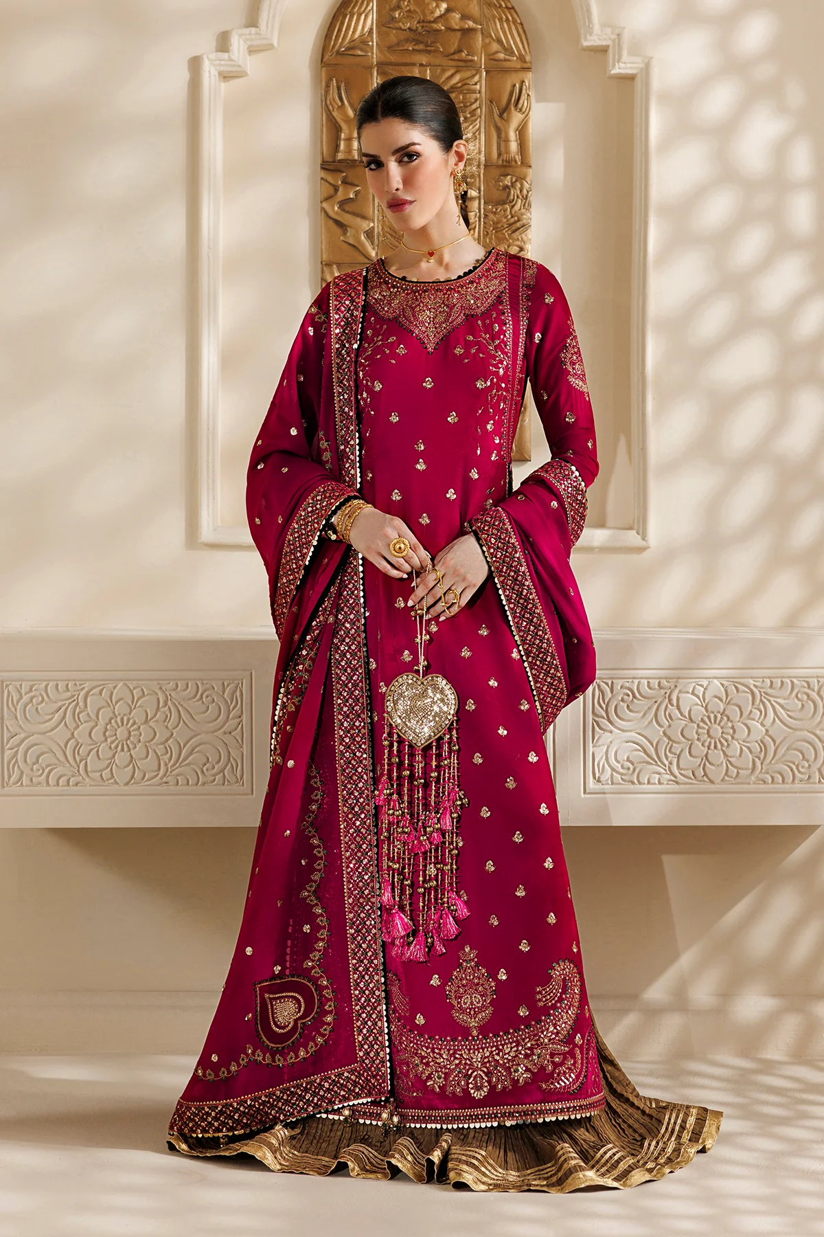 A woman models a traditional deep magenta pink Anarkali gown with intricate gold embroidery, a matching dupatta, and a gold pleated skirt, set against a warm beige background with decorative arches and soft shadow overlays.