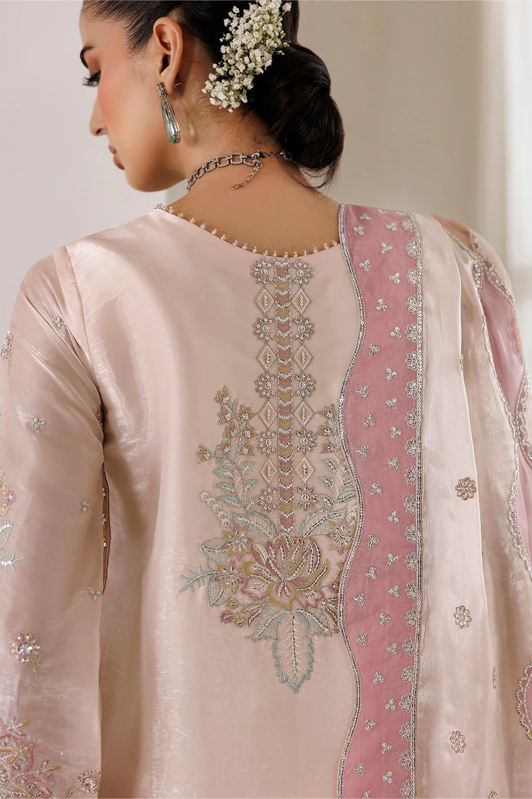 A close-up of a woman wearing a light pink traditional outfit with detailed silver and gold embroidery on the back, featuring a matching pink embroidered dupatta.