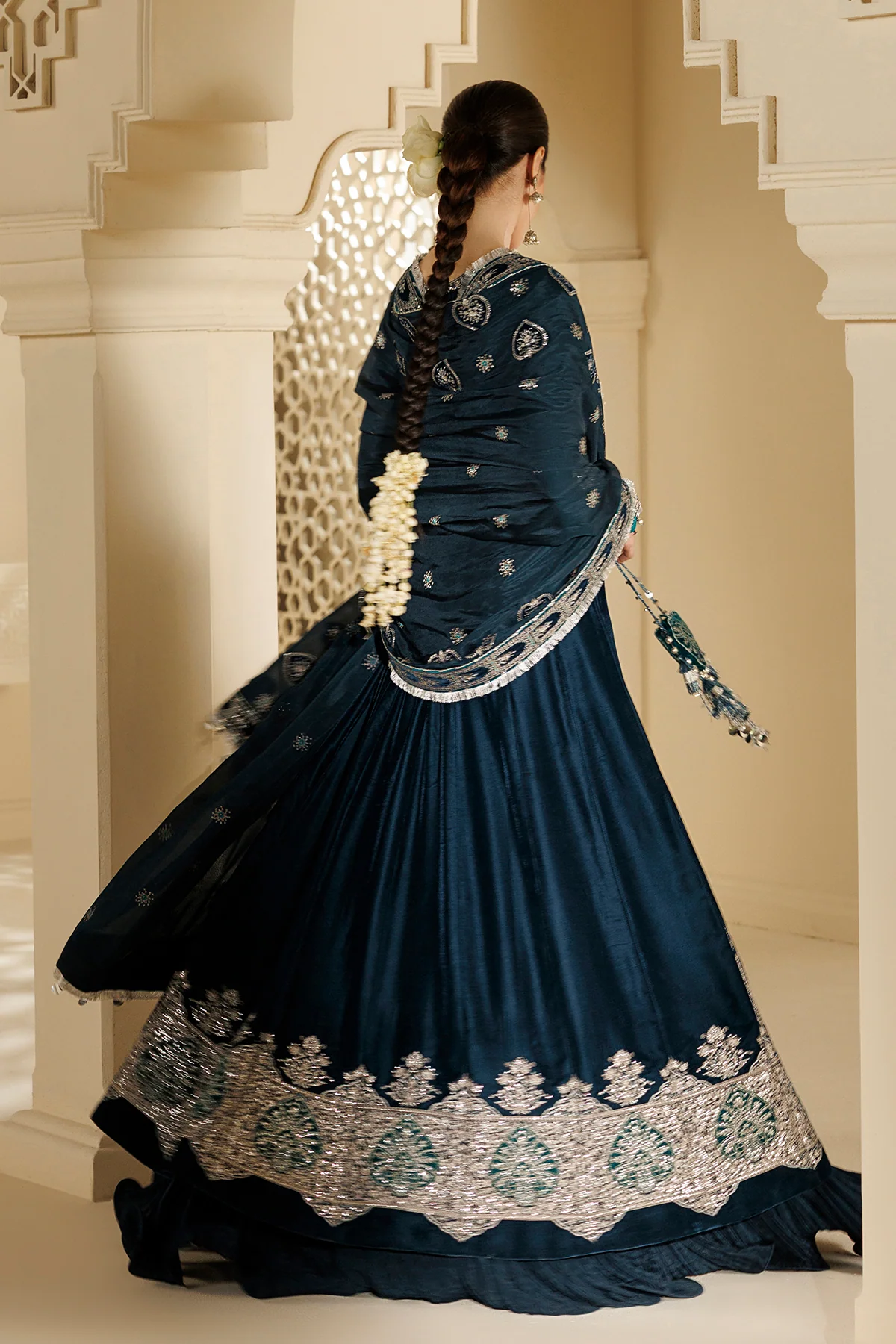 A woman wearing a dark teal blue traditional Indian salwar kameez outfit with intricate silver and white embroidery, shown from behind in an elegant architectural setting.