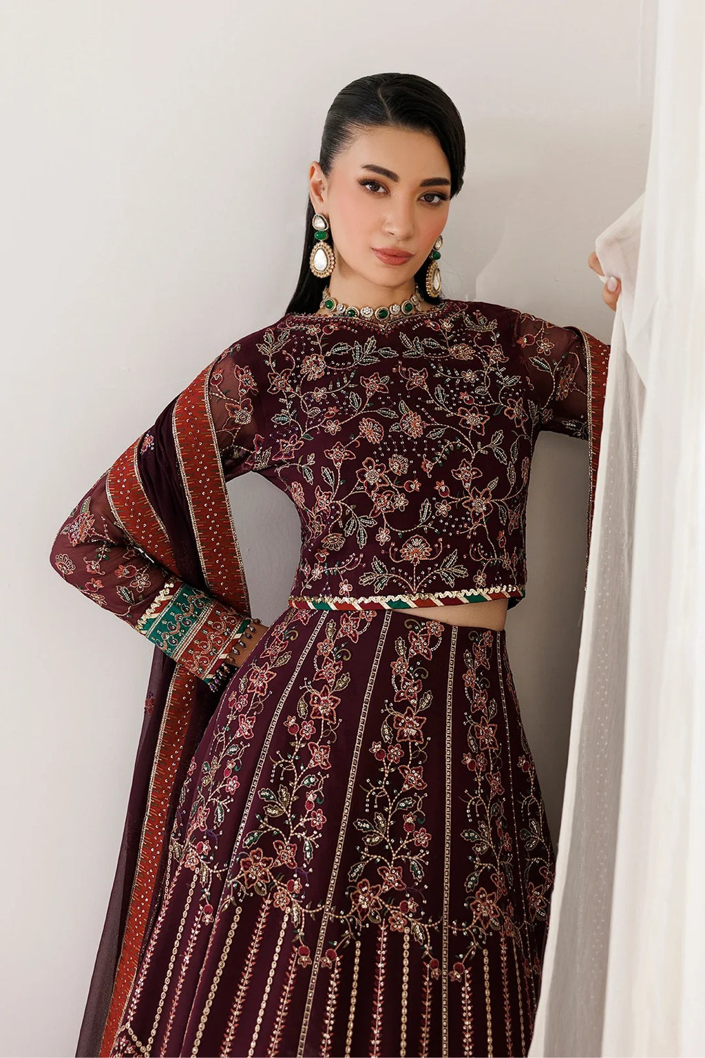 A woman models a traditional embroidered Indian outfit, featuring a deep maroon velvet kurta and matching skirt with intricate floral embroidery, paired with a coordinating dupatta, set against a neutral indoor background.