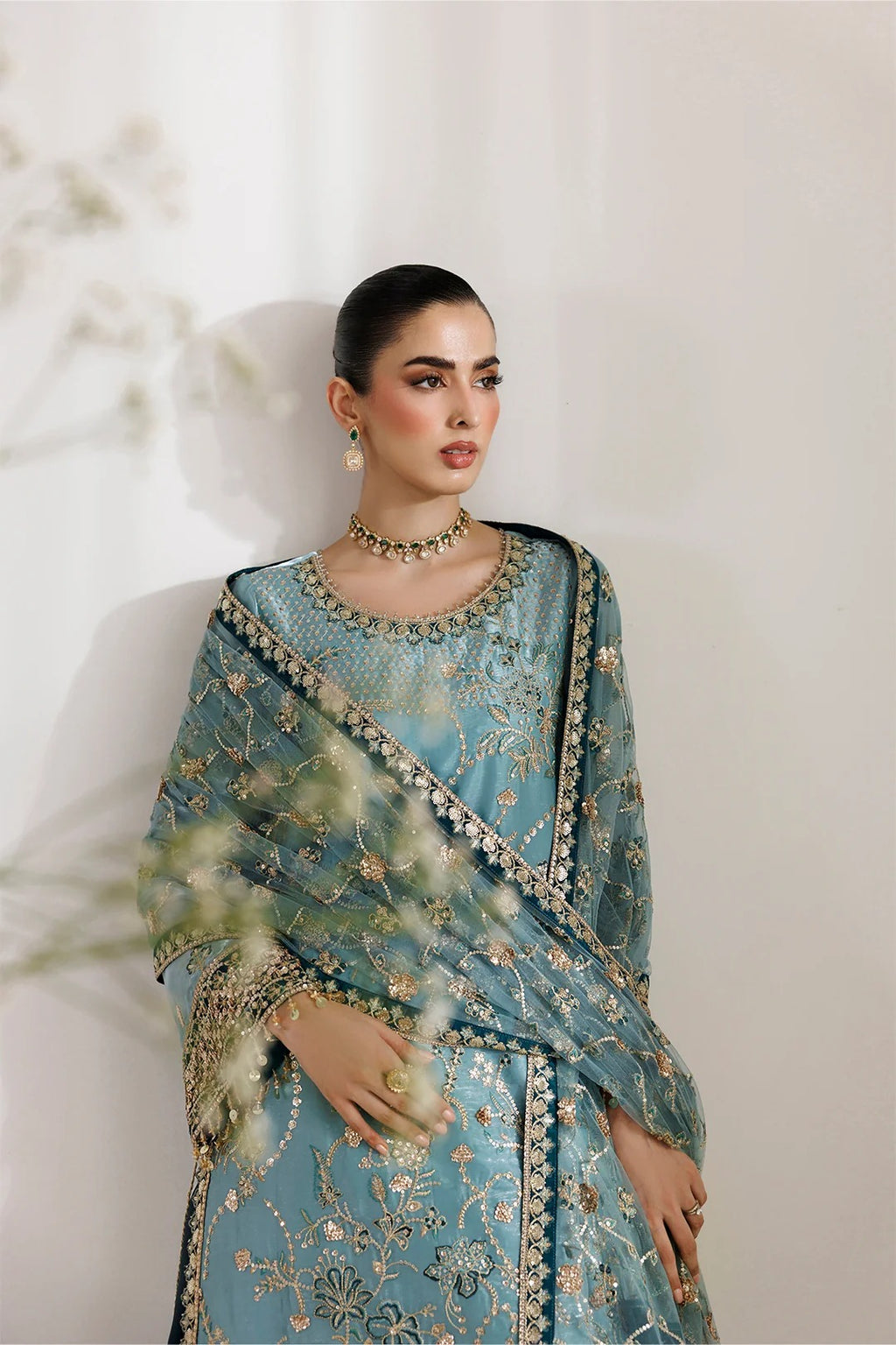 A woman models a traditional light blue embroidered outfit with a matching dupatta, featuring intricate gold and silver floral embroidery, set against a soft beige background.