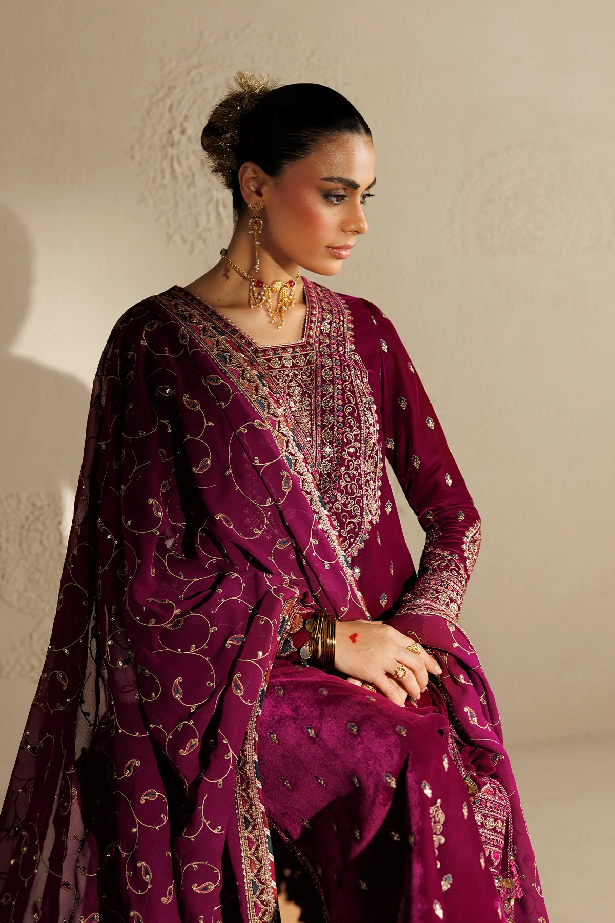 A woman seated and wearing an ornate deep magenta traditional outfit with heavy gold embroidery, a matching embroidered dupatta, and gold jewelry against a warm beige wall.