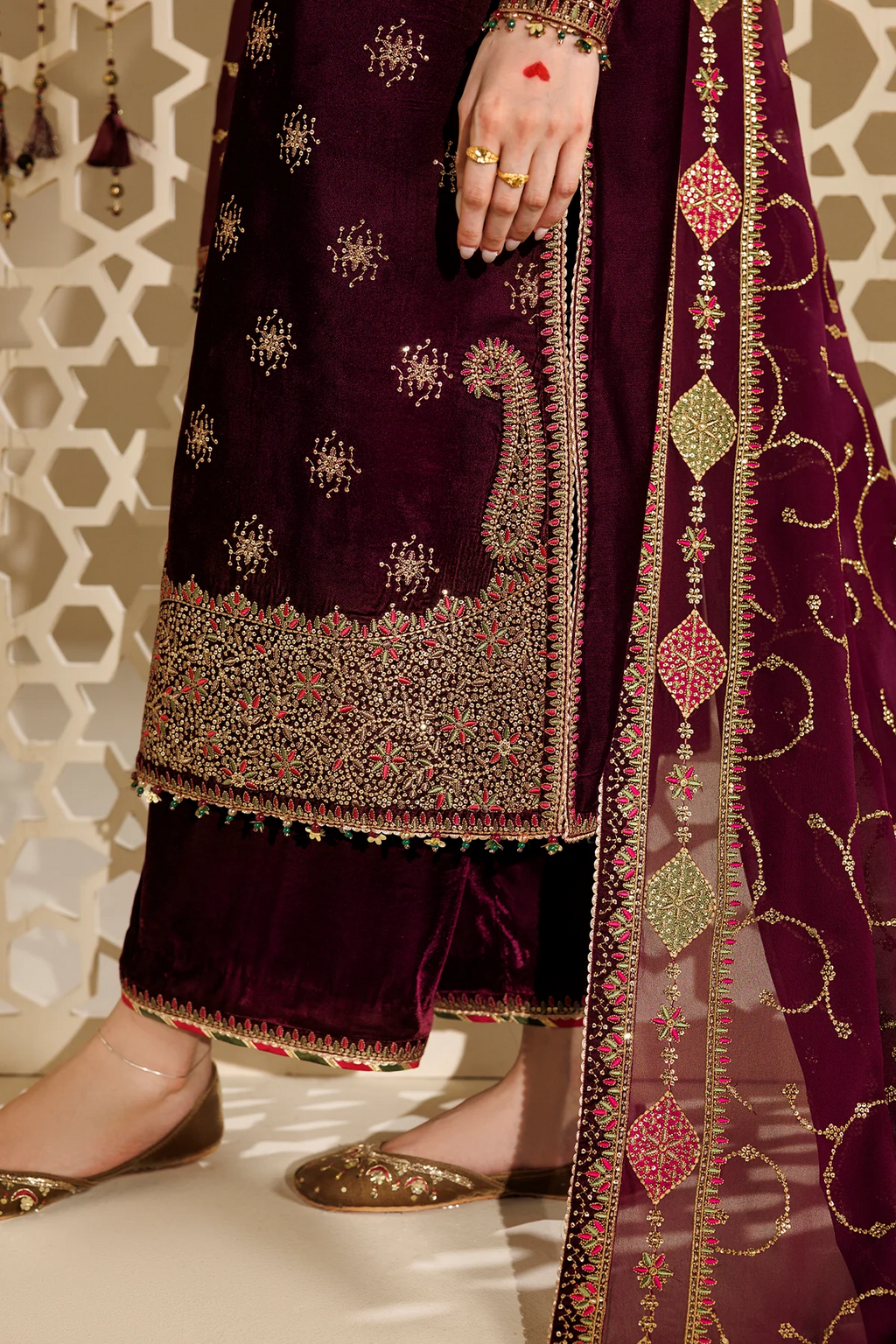 A close-up of a person wearing an ornate deep purple traditional outfit with intricate gold embroidery, a matching sheer dupatta, and flat metallic shoes, standing indoors against a patterned beige wall.