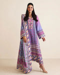 A woman modeling a long-sleeved, floor-length printed kurta in purple, pink, and blue with intricate patterns and borders, set against a plain beige studio background.