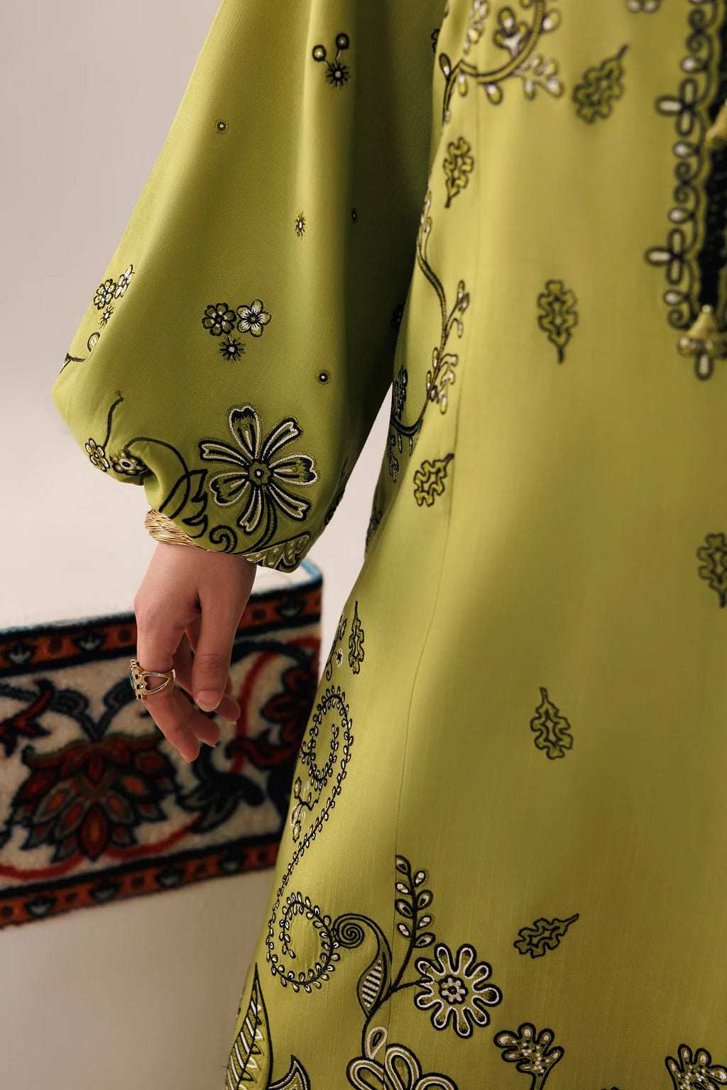 A close-up of a person wearing a flowing olive green tunic or dress with black and white floral embroidery, showing the sleeve, hand with rings, and part of the garment drape.