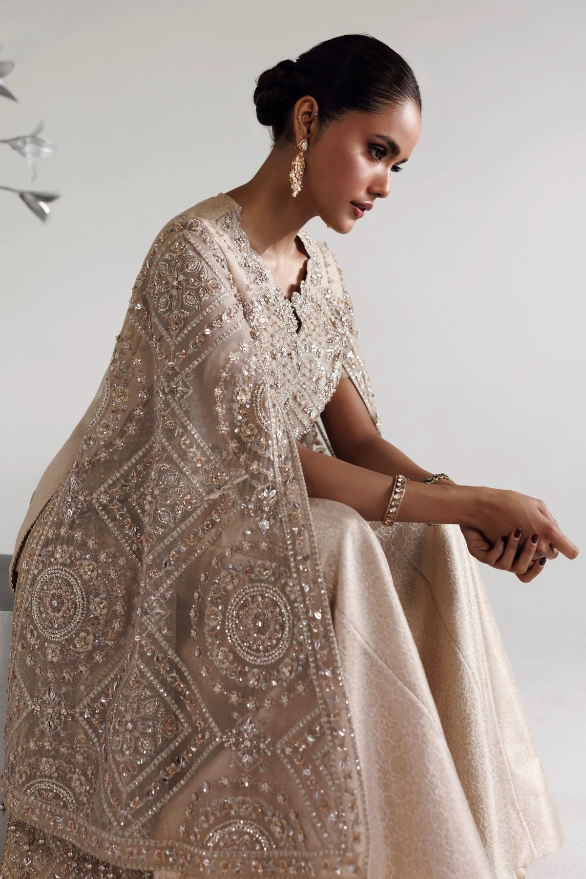 A woman seated in a studio wearing an ornate beige traditional outfit with heavy silver embroidery and beadwork, paired with a matching draped shawl. She is posed against a plain light background, looking downward.