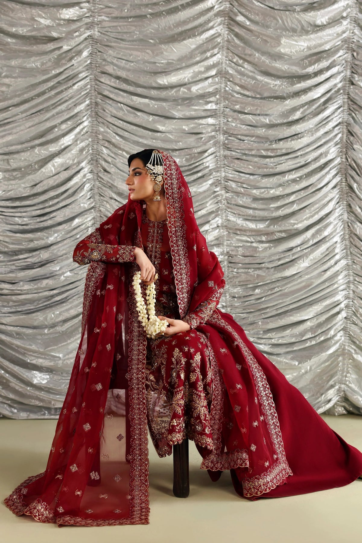 A woman seated on a stool wearing an ornate deep red traditional outfit with heavy gold embroidery, a matching veil, and a white floral garland, posed against a silver draped backdrop.