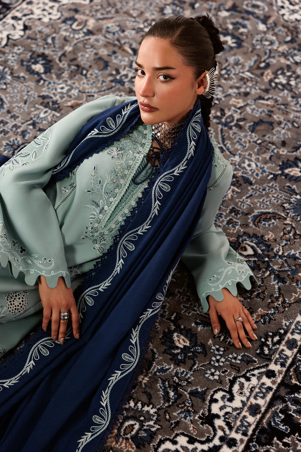 A woman wearing a traditional embroidered outfit with a mint green top and a navy blue draped scarf featuring white floral embroidery, sitting on a patterned rug.