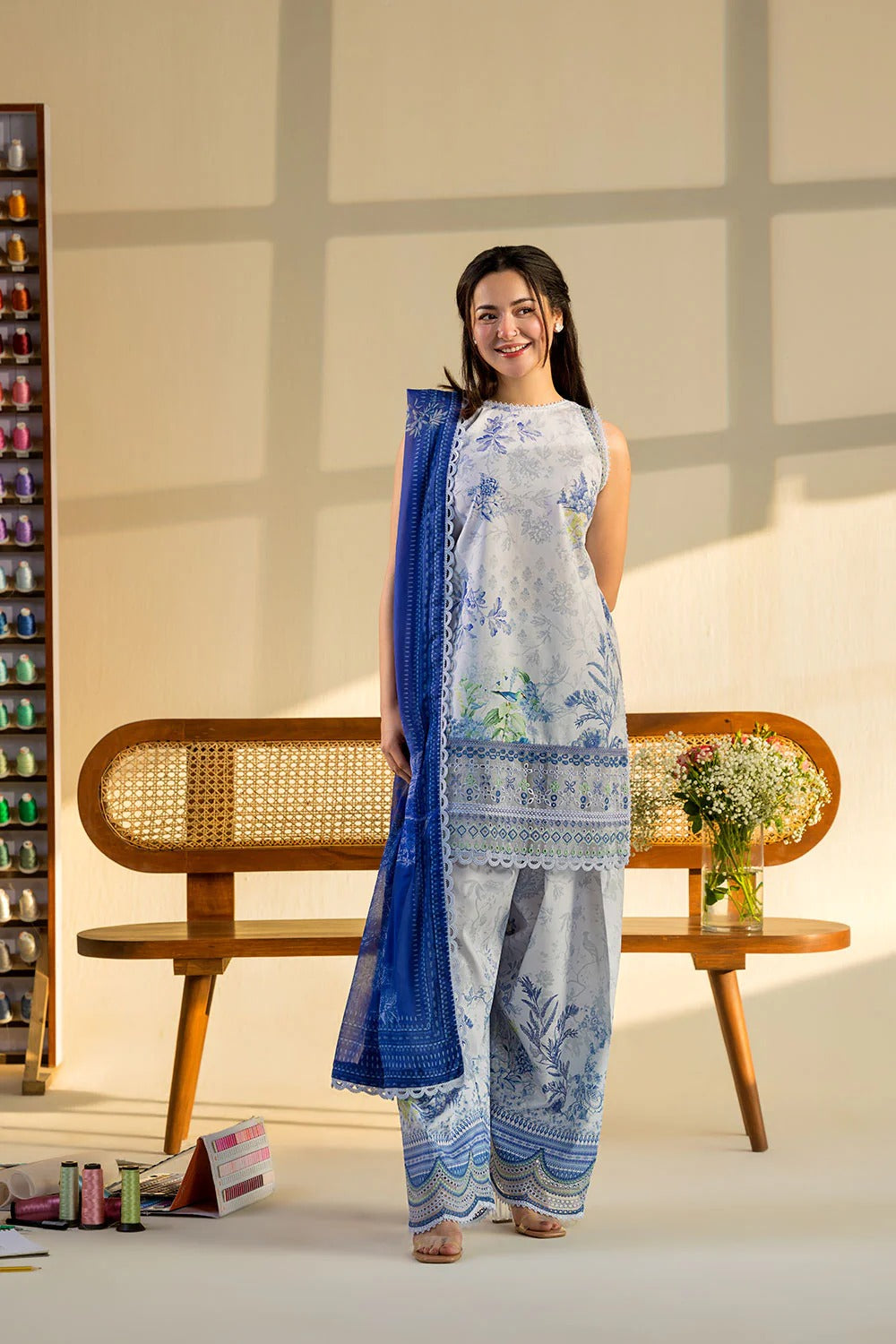 Woman in a blue and white traditional outfit standing in a well-lit room with a wooden bench and decorative items.