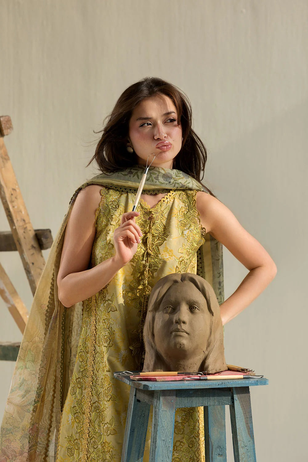 Woman in a green dress holding a paintbrush next to a clay sculpture of a head on a stool.