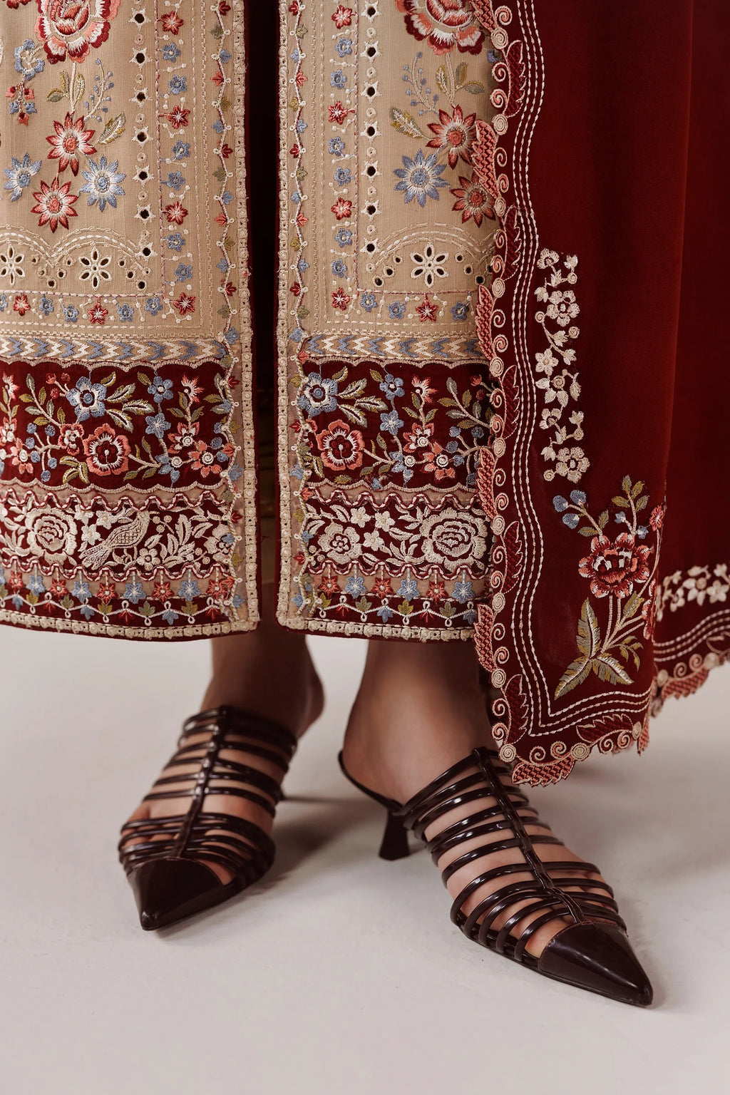 Close-up of a person wearing ornate embroidered traditional clothing with a deep red border and beige embroidered panels, paired with dark brown strappy pointed-toe heeled sandals.