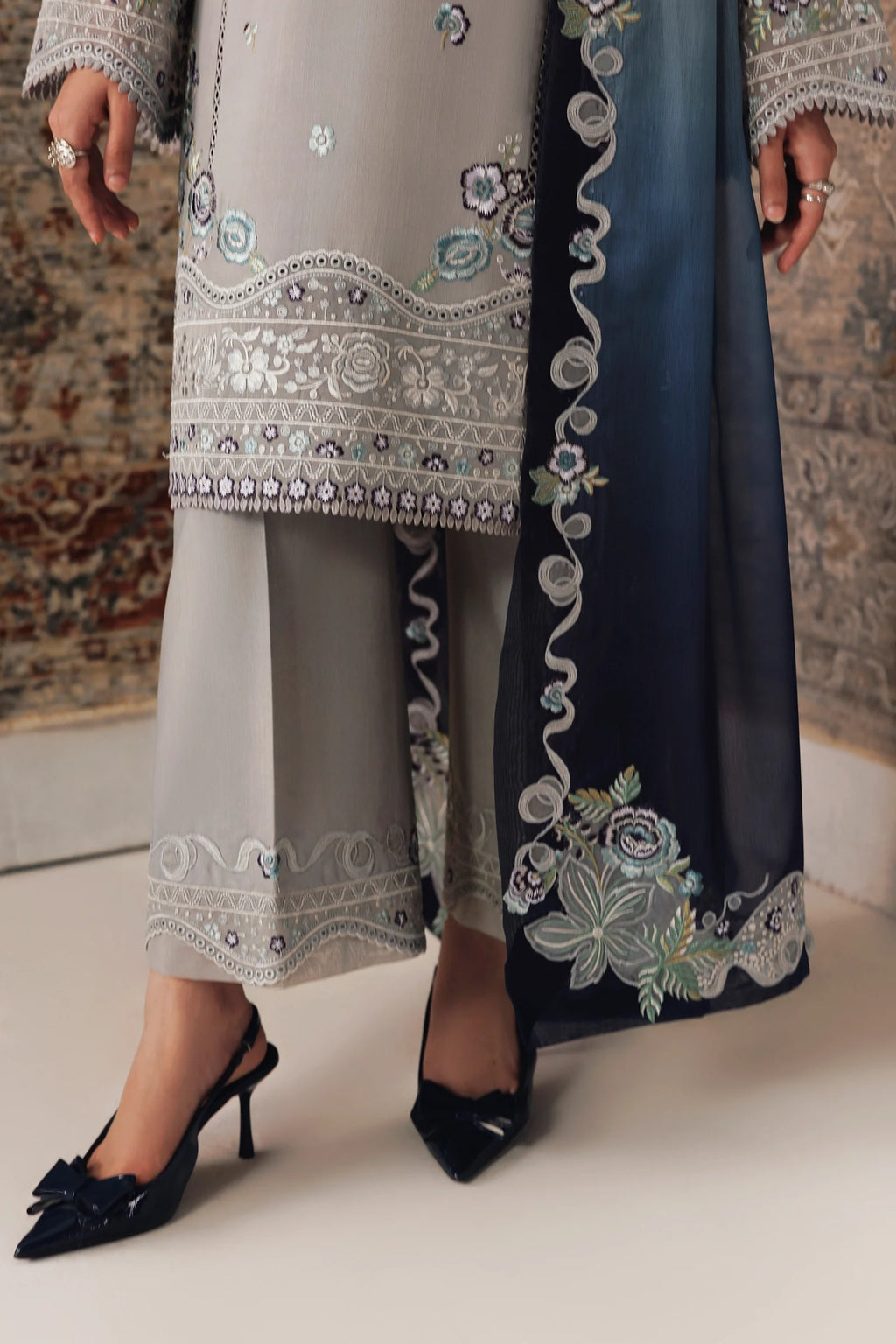 A close-up of a person wearing an embroidered traditional outfit with a light gray tunic and wide-leg pants, a dark navy dupatta with floral embroidery, and black pointed heels.