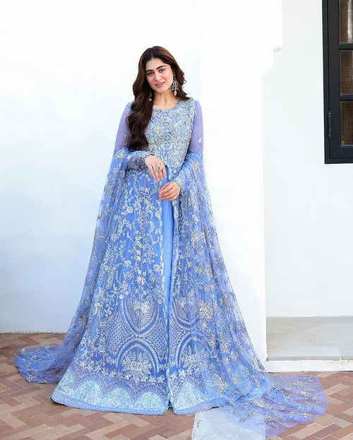 A woman models a light blue embroidered traditional gown with sheer sleeves and a long train, standing against a white wall.