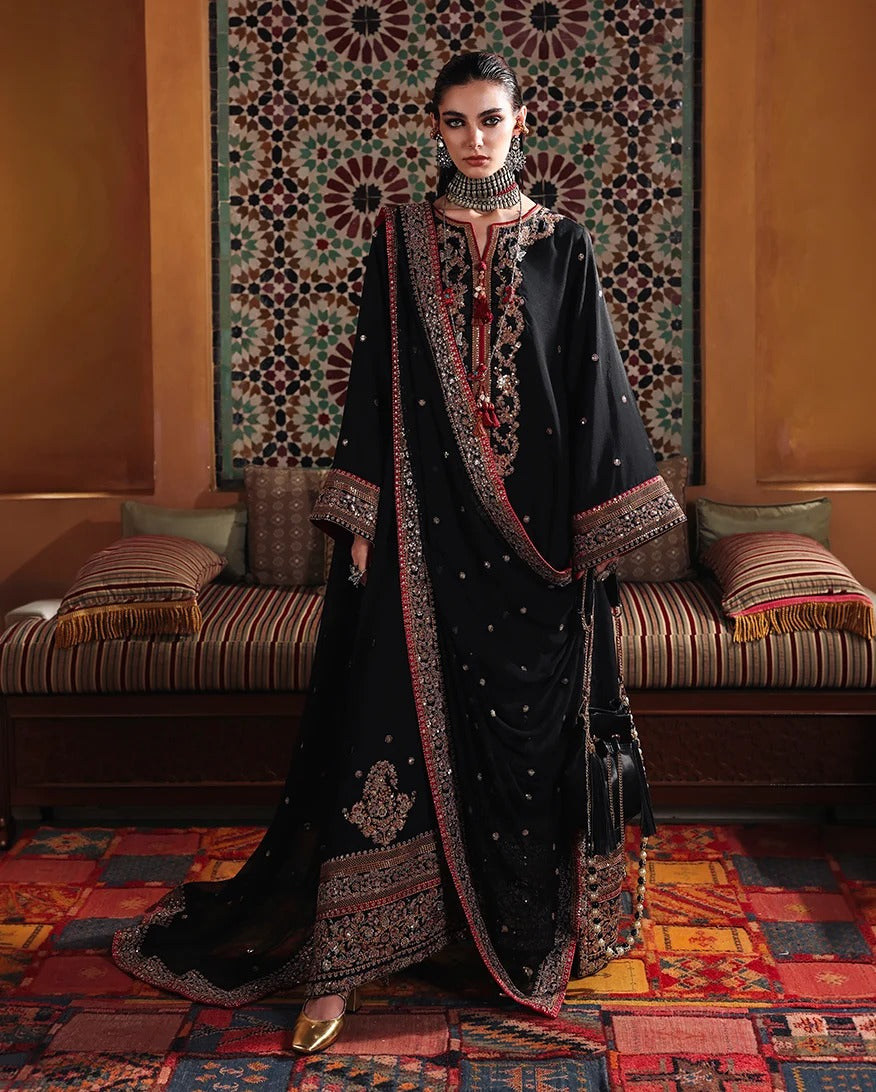 A woman models a traditional black embroidered outfit with a matching dupatta in an ornate room with patterned rugs and a decorative wall hanging.