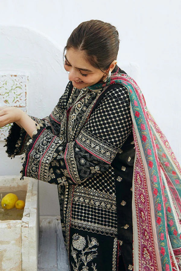 A woman wearing a black three-piece embroidered suit with intricate silver and white floral patterns and a multicolored pink, green, and white border dupatta, standing next to a stone basin containing lemons.