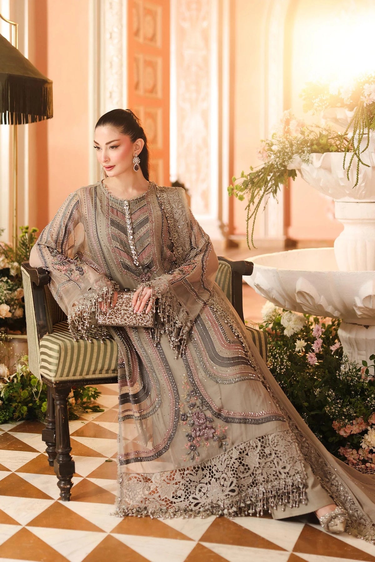 A woman models a traditional South Asian salwar kameez suit in a warm, ornate indoor setting. The outfit is a taupe-beige sheer mesh with intricate multicolor embroidery, geometric patterns, and a matching dupatta draped over her lap.