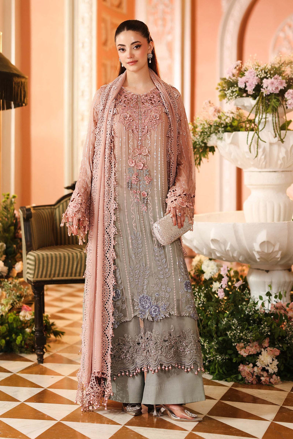 A woman models an elegant, heavily embroidered traditional South Asian outfit in a warm, ornate indoor setting. The outfit consists of a light gray-green kurta with pink and purple floral embroidery, paired with a matching dusty pink dupatta and a sheer pink jacket.