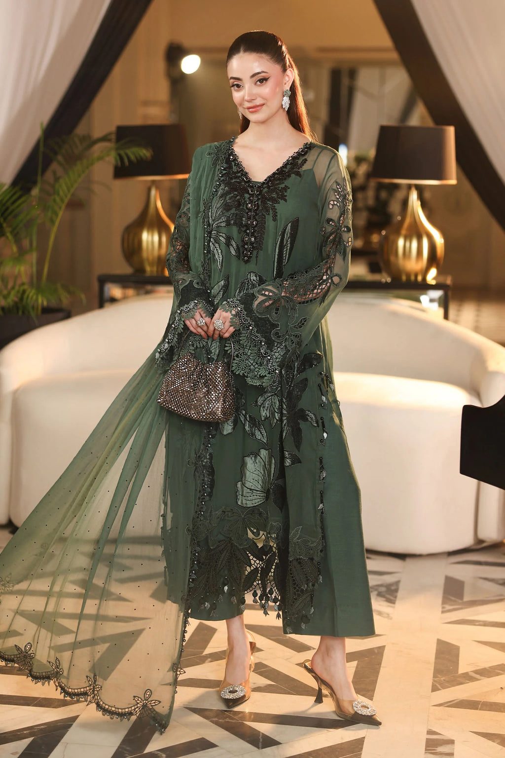 A woman models a dark green embroidered traditional outfit with sheer sleeves and a matching dupatta in an elegant indoor setting.