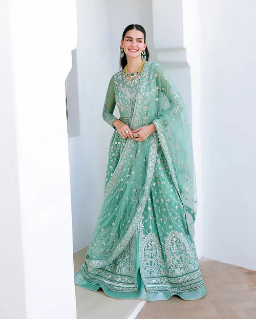 A woman models a traditional mint green Indian ethnic outfit, featuring a heavily embroidered long kurta, matching skirt, and sheer dupatta, set against a bright white architectural background.