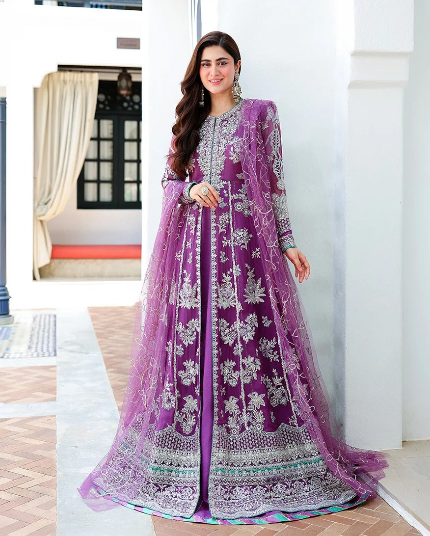 A woman models a traditional purple Anarkali gown with intricate silver embroidery and a matching sheer purple dupatta in an outdoor courtyard setting.