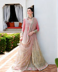 A woman stands outdoors against a white wall wearing an ornate pink and beige embellished traditional outfit with a sheer dupatta. The setting includes a garden bed with pink flowers and a doorway in the background.