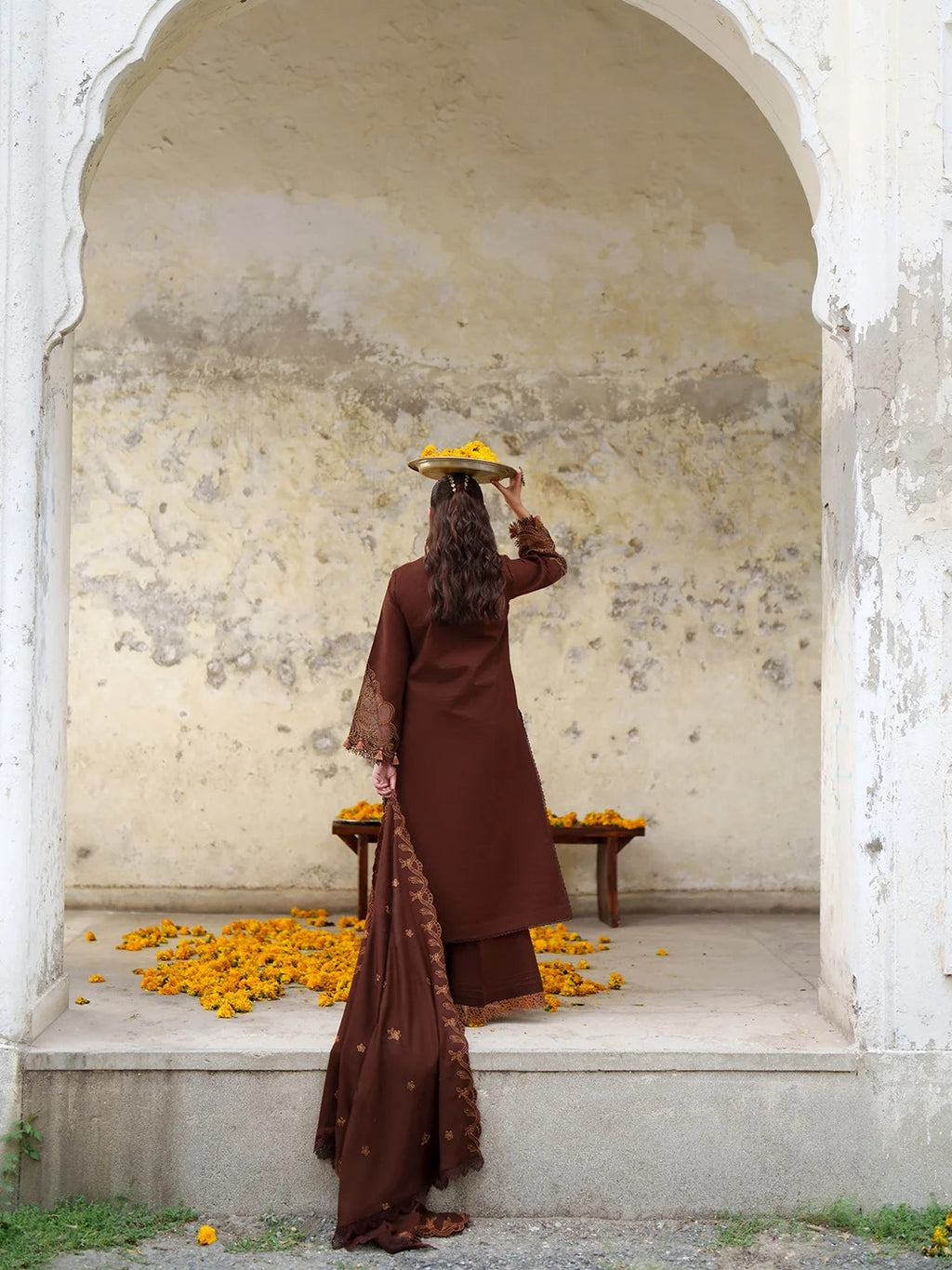 A woman in a brown traditional outfit stands with her back to the camera inside a white arched alcove, holding a small tray of yellow marigold flowers. Yellow flowers are scattered on the floor and a small table.