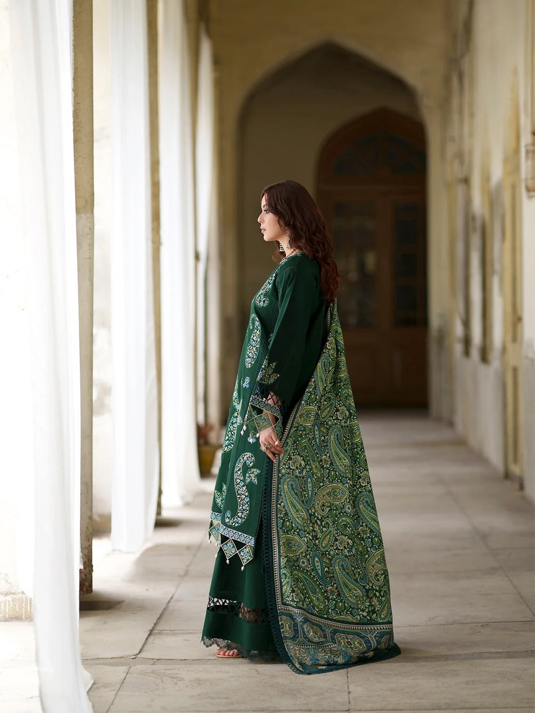 A woman stands in profile in a sunlit corridor wearing a dark green traditional outfit with ornate gold and silver embroidery and a matching patterned dupatta.