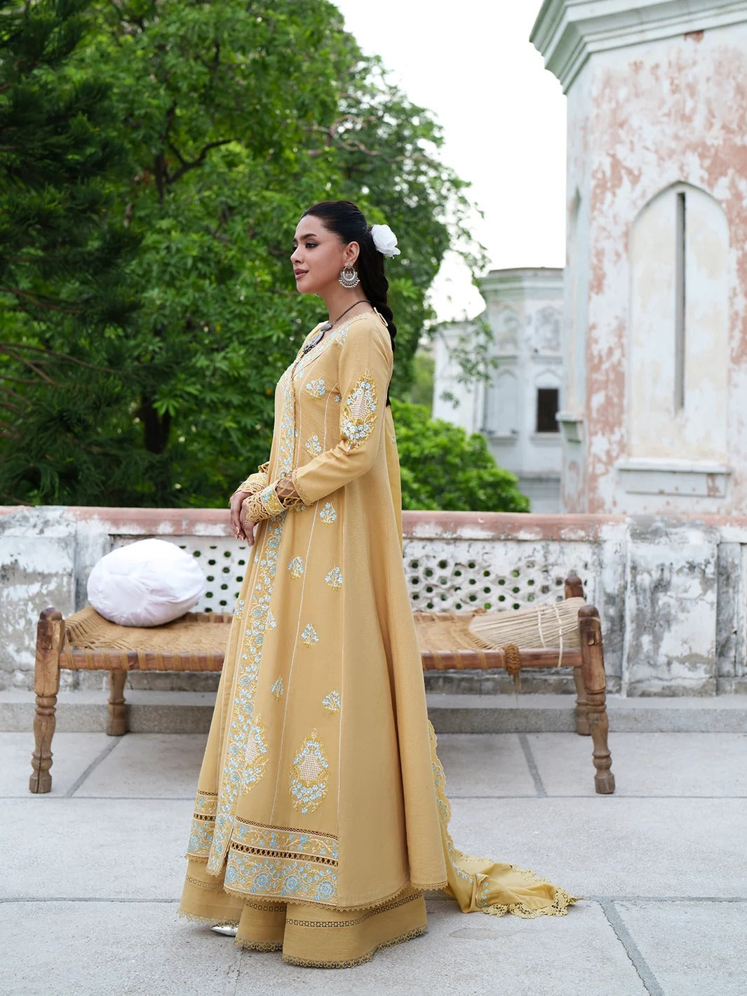 A person wearing a long yellow embroidered traditional dress with intricate gold and light blue floral patterns, standing on a stone terrace.