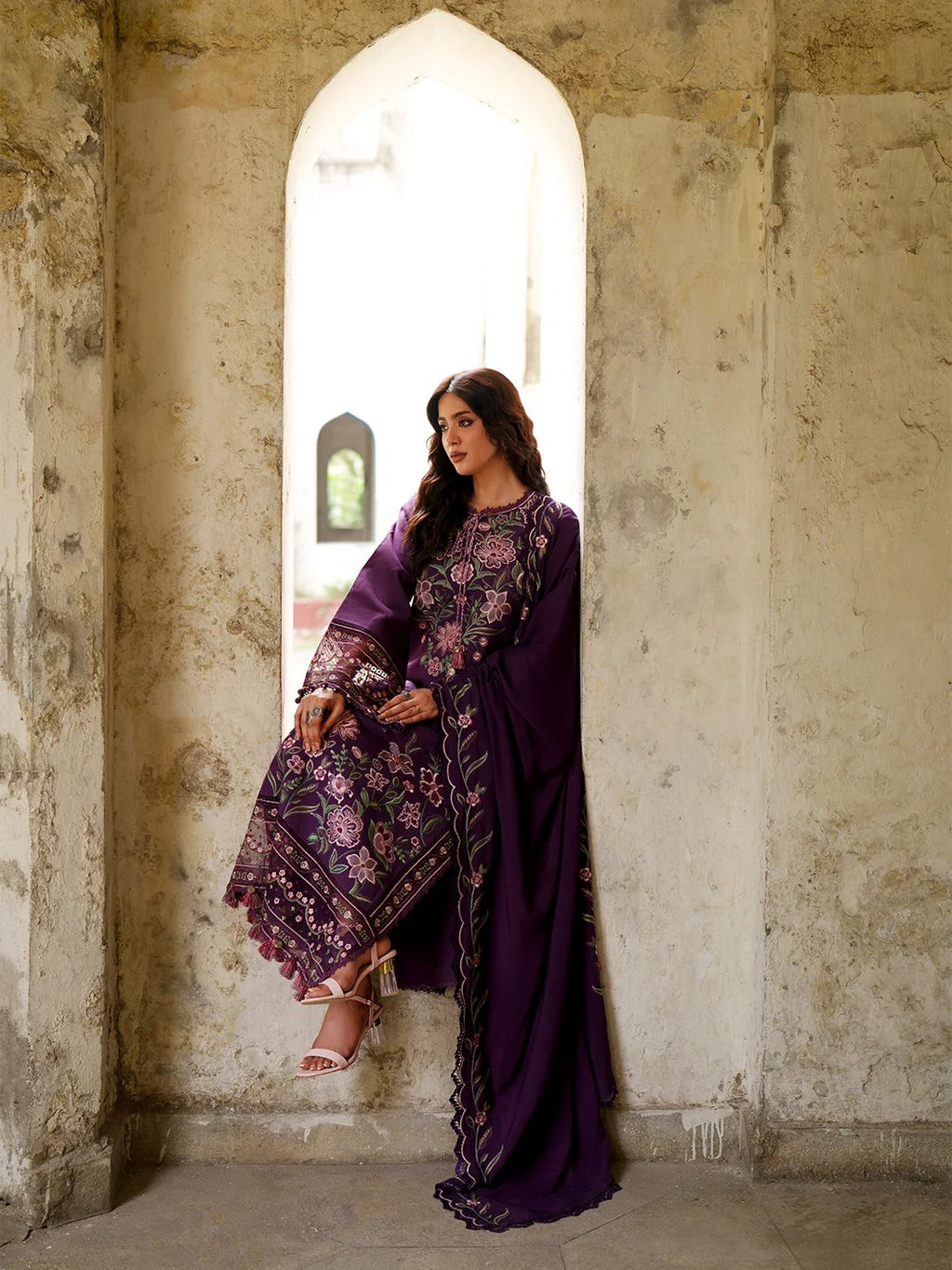 A woman models a deep purple embroidered traditional outfit with floral patterns and a matching dupatta, posed in a rustic stone archway setting.