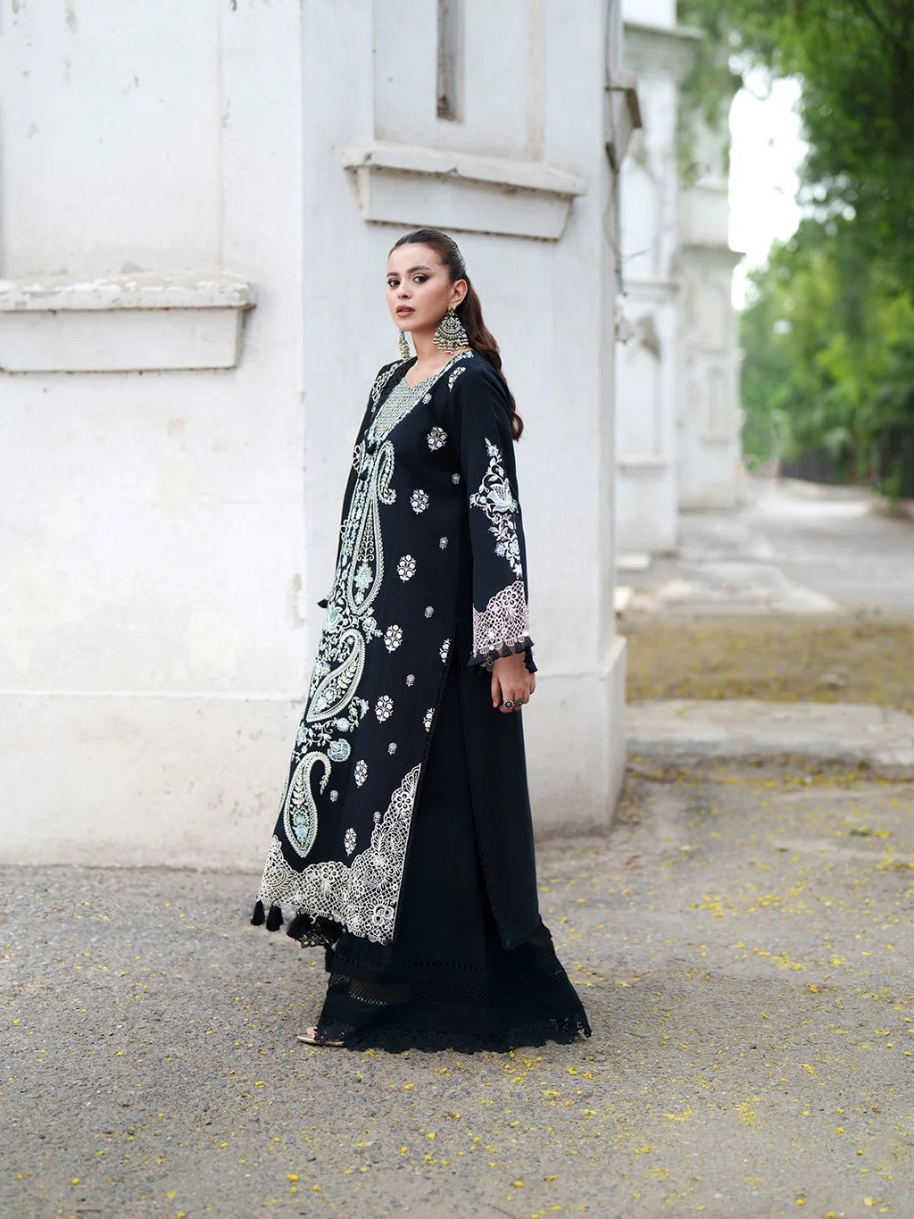 A woman models a long black embroidered dress with intricate silver, white, and light blue paisley patterns, standing outdoors next to a white building.