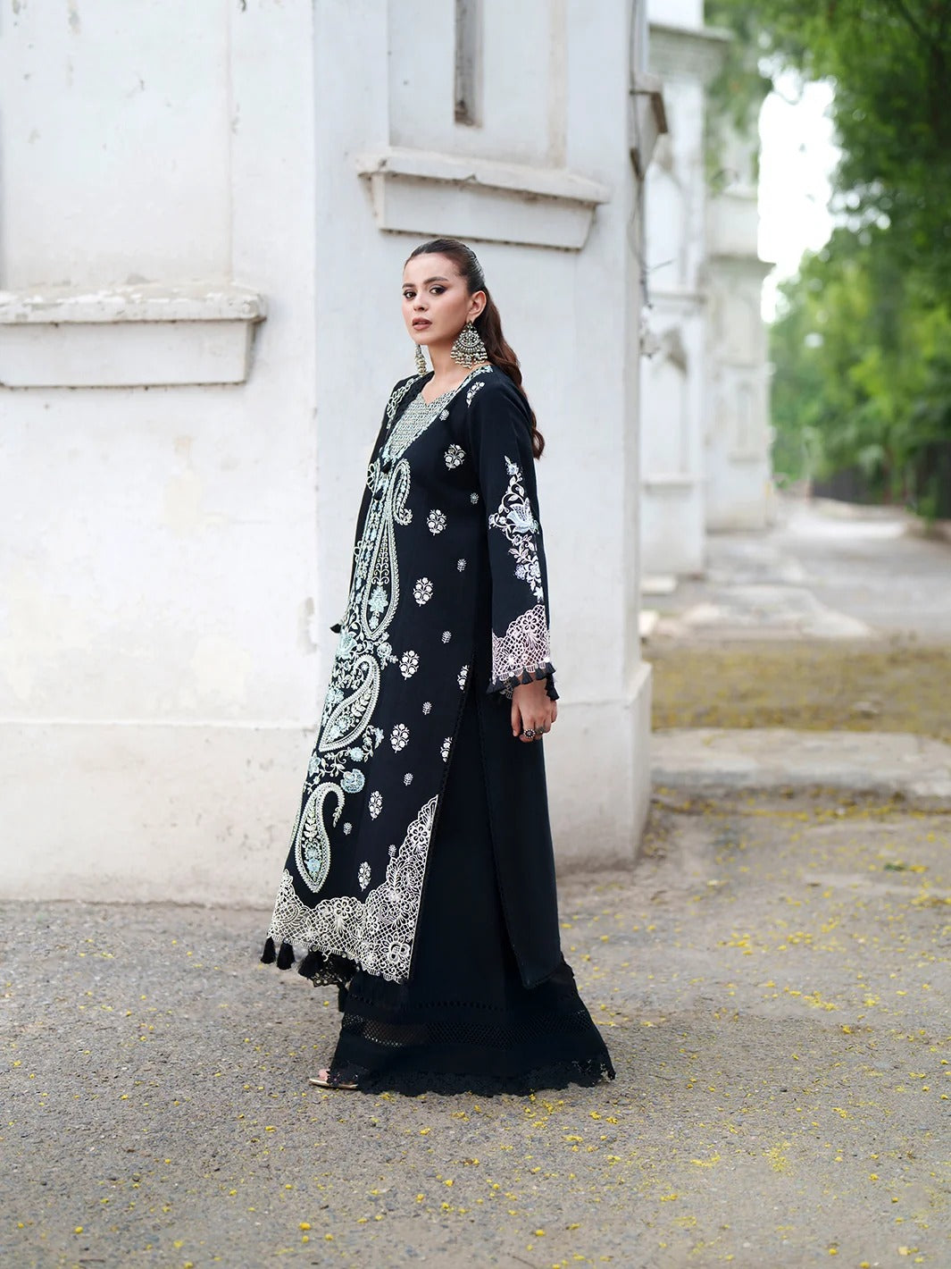 A woman models a long black embroidered dress with intricate silver, white, and light blue paisley patterns, standing outdoors next to a white building.