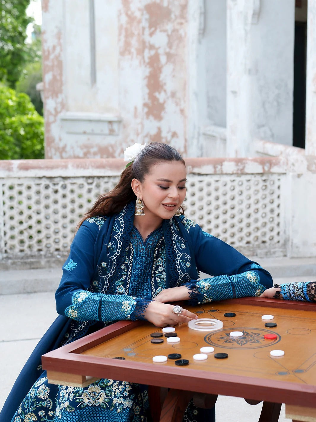 A woman in a blue and white embroidered traditional outfit sits at a wooden table playing a game of Go with black and white stones. The background shows a weathered stone wall and green foliage.